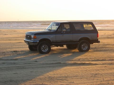 1989 Ford Bronco - A two-tone Ford Bronco sits on a sandy beach with the ocean in the background during sunset.