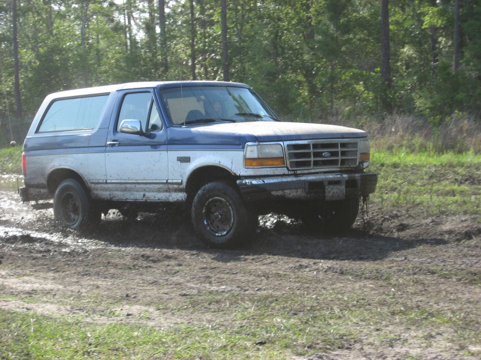 Blue and white Ford Bronco driving through mud. Grille and headlights indicate early 90s model