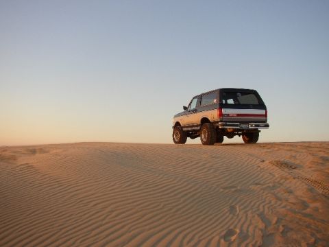 1989 Ford Bronco - A Ford Bronco drives on a sand dune under a clear sky.
