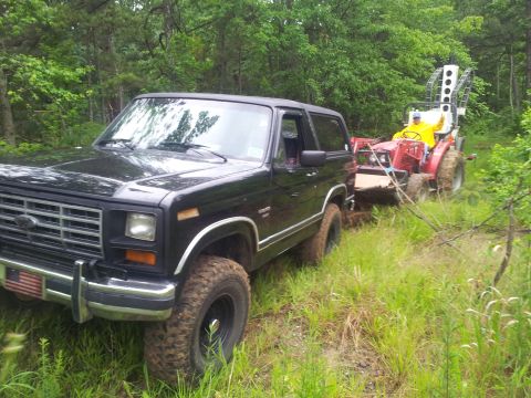 1986 Ford Bronco - A black Ford Bronco tows a red tractor through a grassy, wooded area.