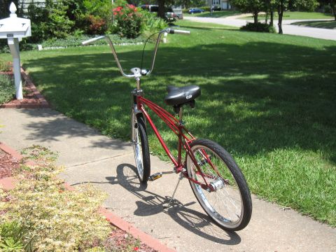 1994 Ford Bronco - A red cruiser bicycle with distinctive high handlebars is parked on a concrete sidewalk next to a grassy lawn.
