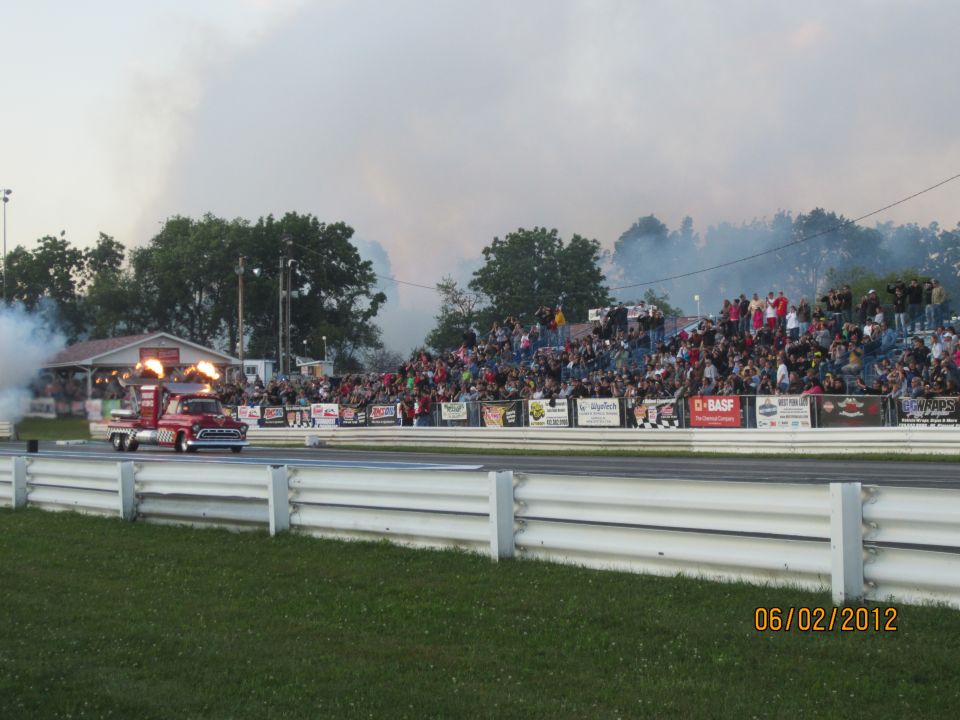 Red pickup truck with flames shooting from exhaust pipes on a drag strip with spectators in the stands