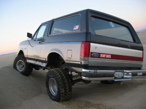 1989 Ford Bronco - This lifted Ford Bronco is shown from the rear, driving on a sandy terrain under a clear sky.