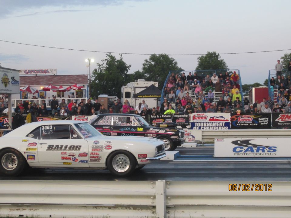 White drag car with 'BALLOG' and sponsor decals, next to a black drag car, on a race track with spectators
