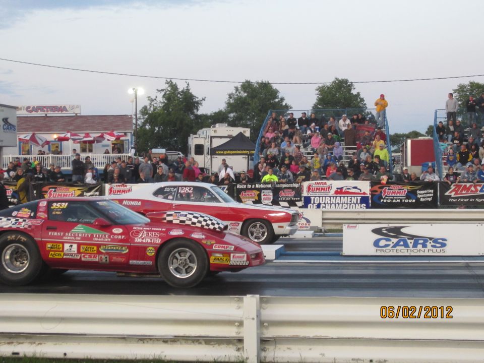 Two drag racing cars, a red Chevrolet Corvette and a red and white muscle car, lined up on a drag strip with spectators in the background