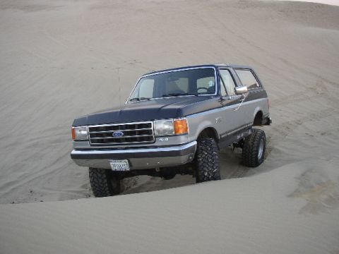 1989 Ford Bronco - A lifted Ford Bronco navigates sandy terrain, its two-tone paint job contrasting with the desert landscape.