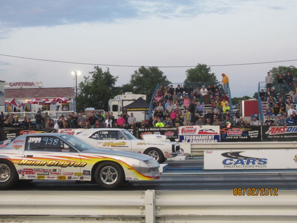 Two drag racing cars lined up on a track, with spectators in the stands. Date stamp reads 06/02/2012