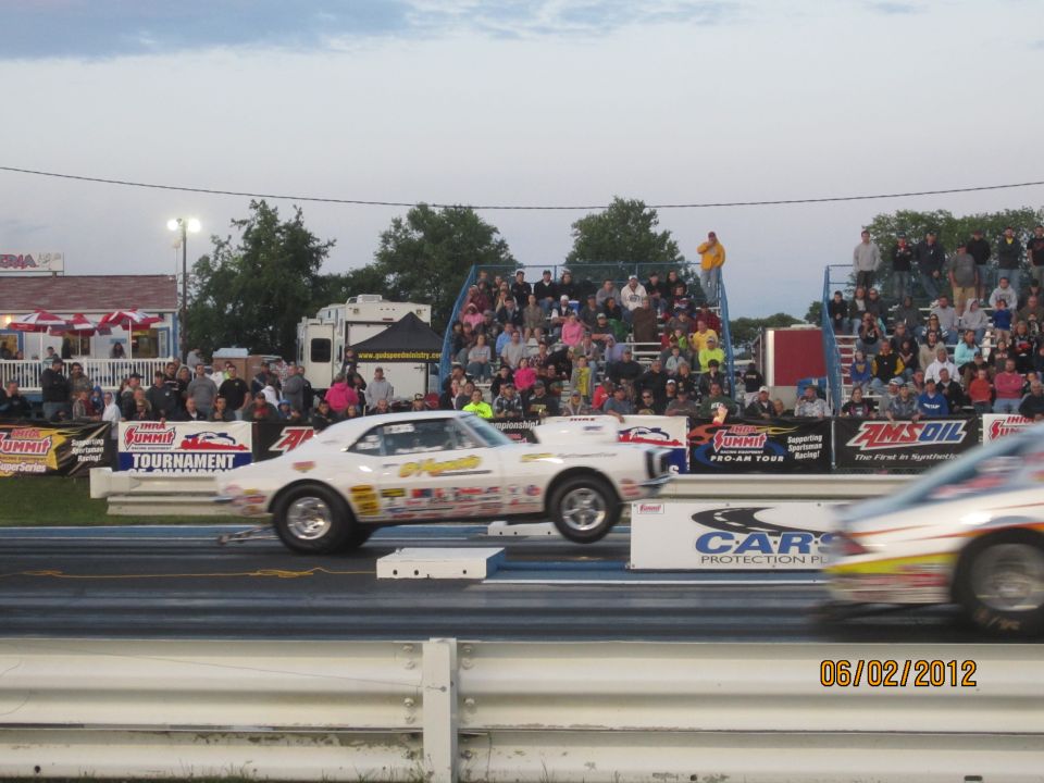Two drag cars race down a track with spectators in the stands. One car is in focus, the other is blurred