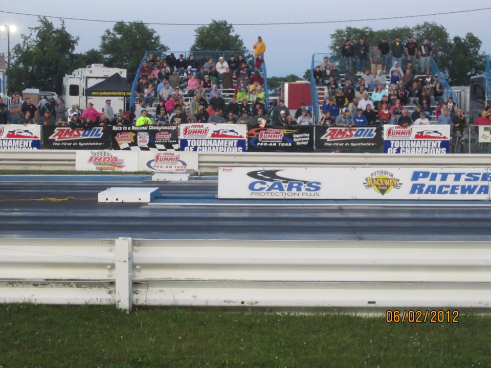Drag strip with bleachers full of spectators and sponsor banners. Date stamp reads 06/02/2012