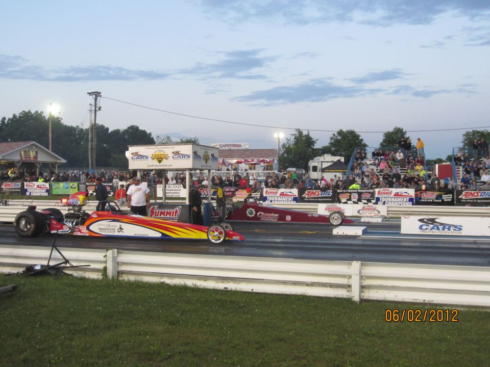 Drag racing cars on a track, one red and one red with yellow and blue flames, with spectators in the background