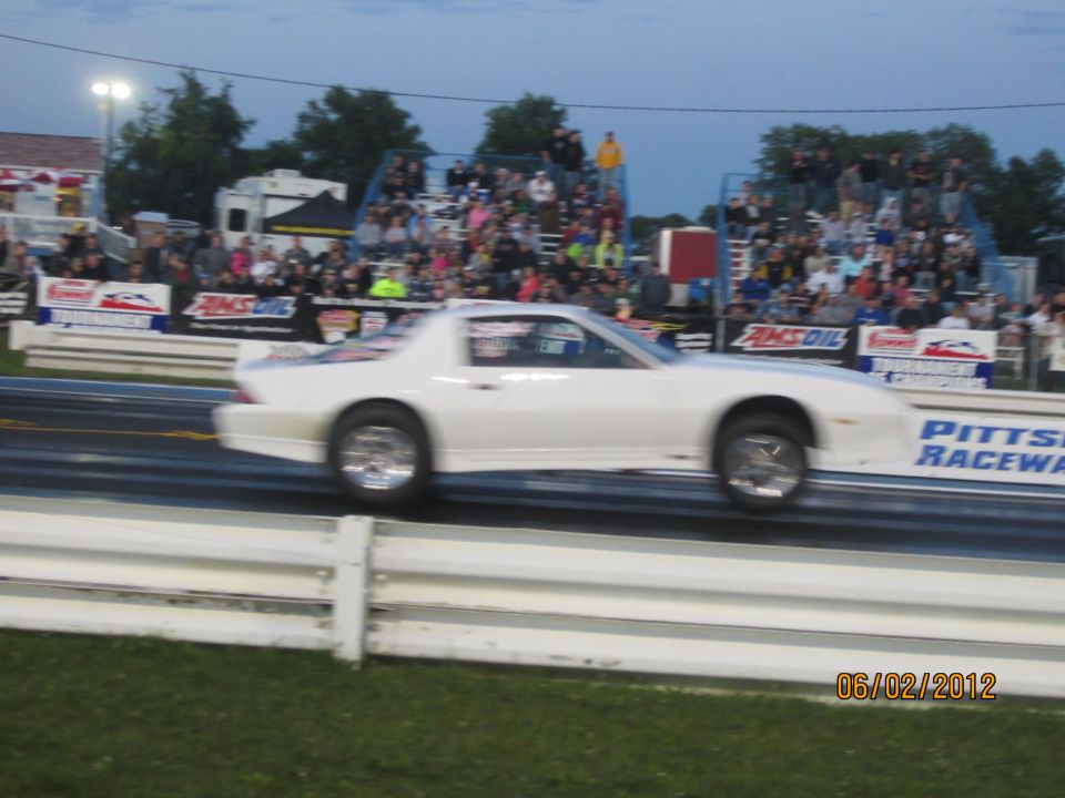 White drag car in motion on a race track, side view, with spectators in bleachers