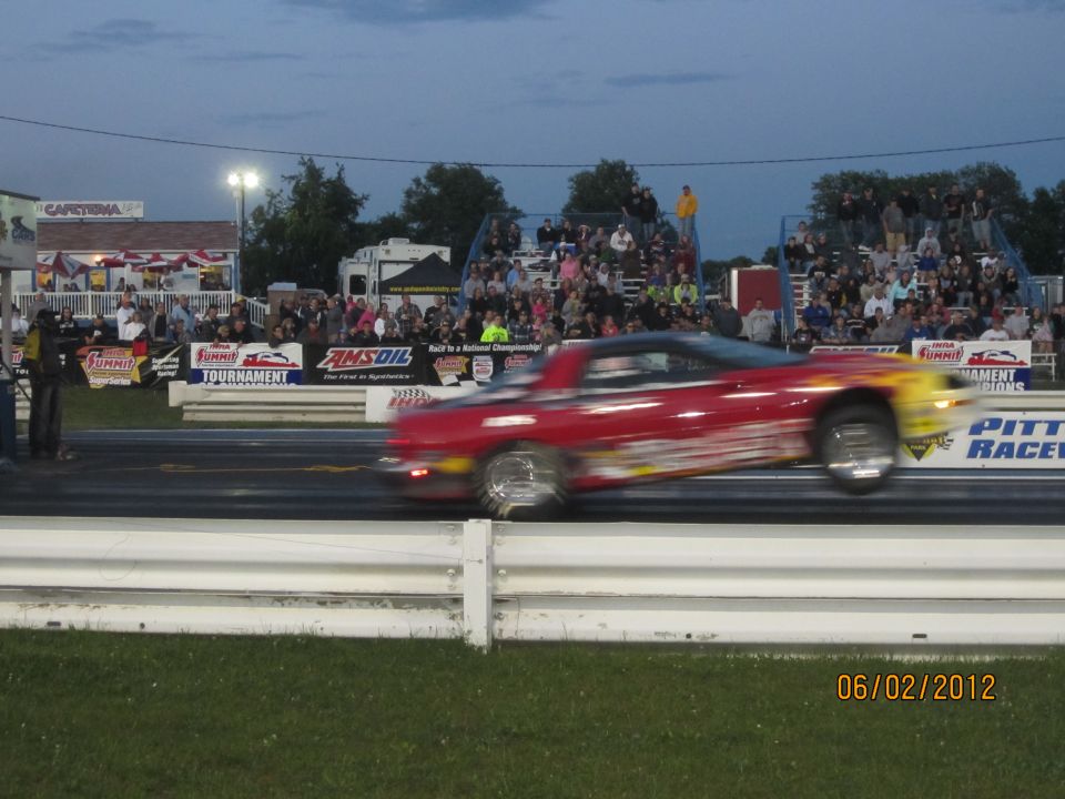 Red drag car with flame decals on a race track, blurred due to motion. Spectators are in the background
