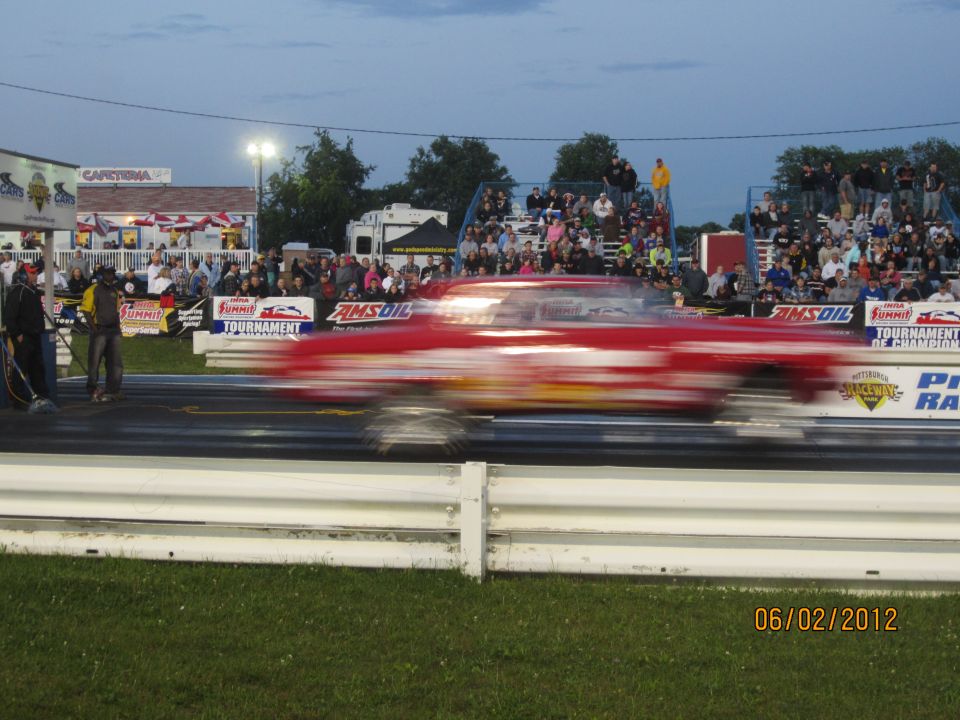 Red drag car speeding down a track with spectators in the background. Date stamp reads 06/02/2012