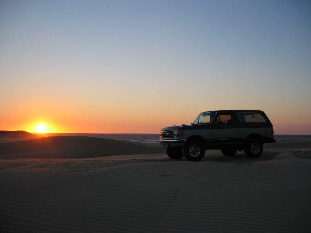 Two-tone Ford Bronco SUV on sand dunes silhouetted against a sunset.