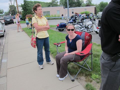 1991 Ford LTD Crown Victoria - Two women on a sidewalk, one standing in a yellow shirt and one seated in a red folding chair, with parked motorcycles behind them.