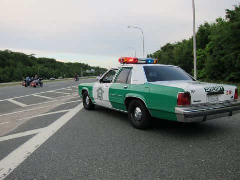 1991 Ford LTD Crown Victoria - green-and-white Ford Sheriff sedan number 351 parked on a highway shoulder as a motorcycle approaches at dusk.