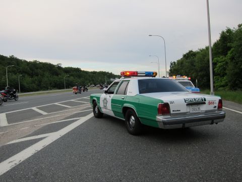 1991 Ford LTD Crown Victoria - Rear three-quarter view of a green-and-white Ford Sheriff sedan on a highway with motorcycles riding in the distance at dusk.
