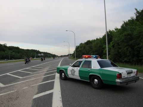 1991 Ford LTD Crown Victoria - green-and-white Ford Sheriff sedan parked diagonally across a multi-lane highway as motorcycles approach in the distance at dusk.