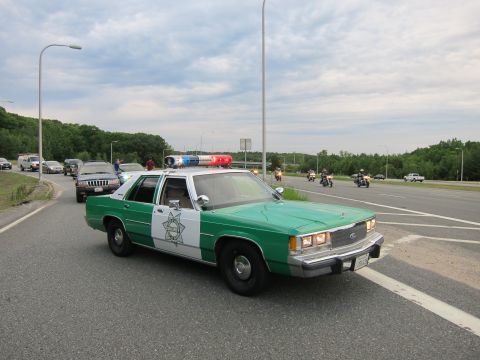 1991 Ford LTD Crown Victoria - green-and-white Ford Sheriff sedan with emergency lights lit leads vehicles and motorcycles along a highway at dusk.