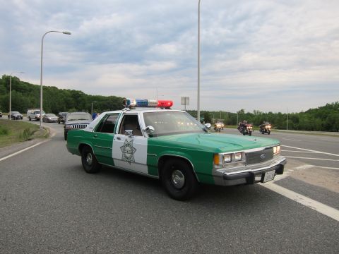 1991 Ford LTD Crown Victoria - green-and-white Ford Sheriff cruiser with its light bar activated sits on a highway as motorcycles ride past at dusk.