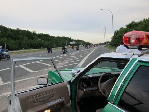 1991 Ford LTD Crown Victoria - View from inside a green police car with both front doors open, looking out at motorcycles riding along the highway at dusk.