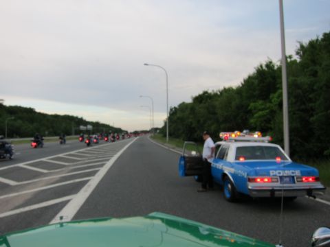 1991 Ford LTD Crown Victoria - blue Police cruiser with emergency lights and open door parked on a highway shoulder as a line of motorcycles rides by at dusk.