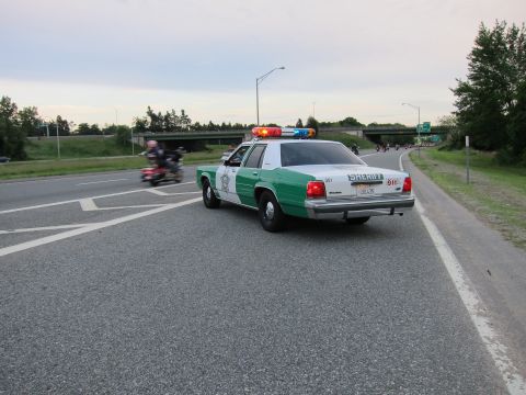 1991 Ford LTD Crown Victoria - green-and-white Ford police-style sedan marked SHERIFF sits on a highway with its light bar lit as motorcycles ride past at dusk.