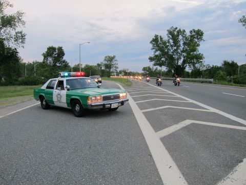 1991 Ford LTD Crown Victoria - green-and-white Ford sheriff sedan with emergency lights activated escorts a line of approaching motorcycles along a divided highway.