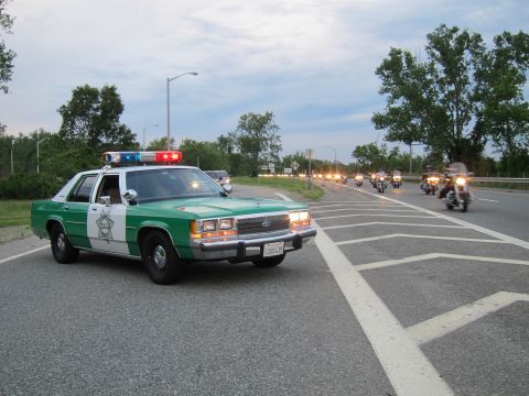 1991 Ford LTD Crown Victoria - green-and-white Ford sheriff sedan runs its light bar on a highway as a group of motorcycles rides toward the camera.