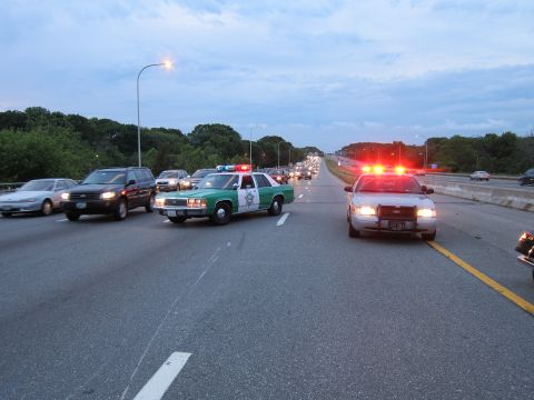 1991 Ford LTD Crown Victoria - green-and-white sheriff sedan and a second patrol car with red lights flashing block lanes of a multi-lane highway among traffic at dusk.