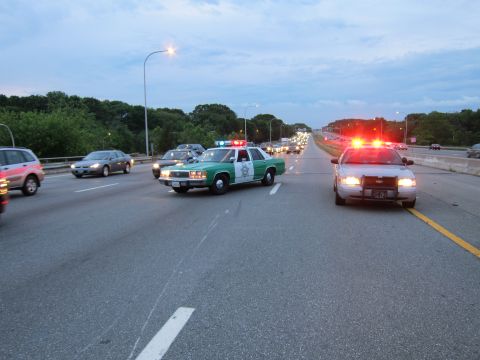 1991 Ford LTD Crown Victoria - green-and-white sheriff sedan and a patrol car with red emergency lights occupy a multi-lane highway alongside passing traffic at dusk.