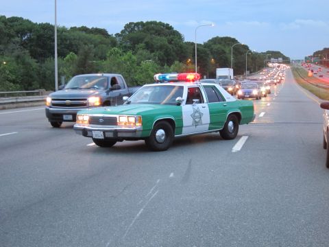 1991 Ford LTD Crown Victoria - green-and-white Ford sheriff sedan runs its light bar on a highway with a pickup truck and heavy traffic backed up behind it at dusk.