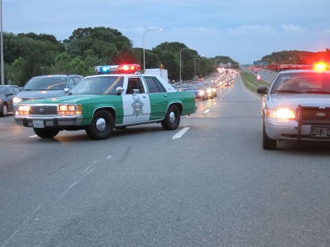 1991 Ford LTD Crown Victoria - green-and-white sheriff sedan and a second patrol car with flashing lights hold a highway lane as a line of traffic waits behind them.