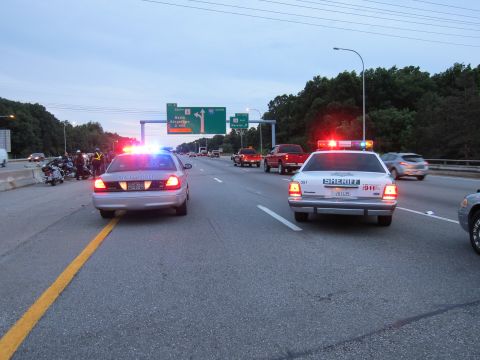 1991 Ford LTD Crown Victoria - Two patrol cars including one marked SHERIFF run emergency lights on a highway near a green overhead road sign, with motorcycles parked on…