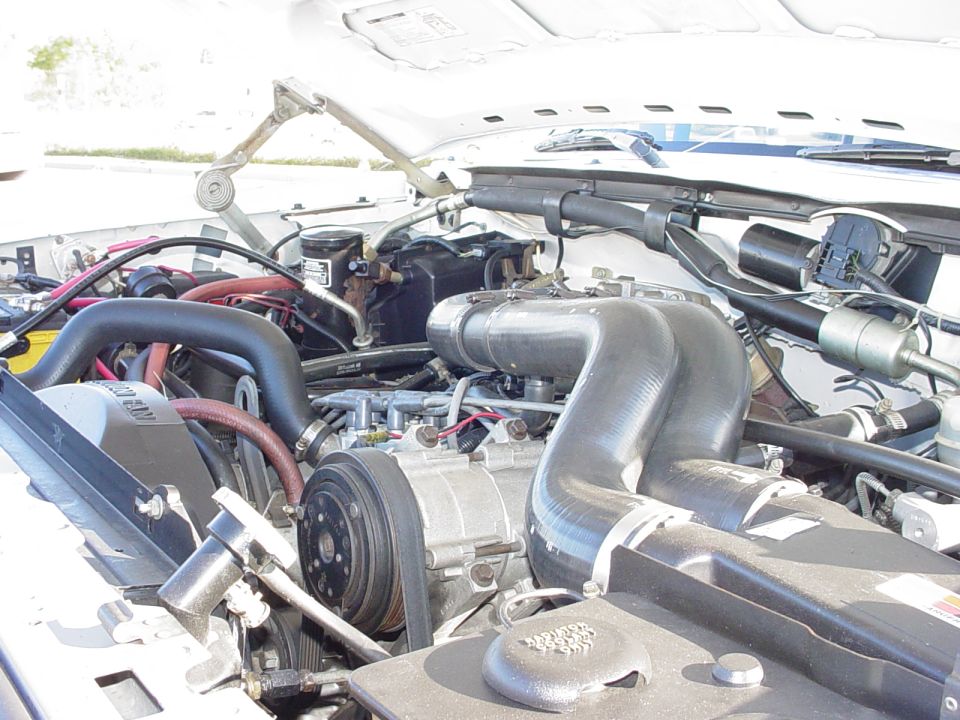 Engine bay of a white Ford Bronco with large black intake hose, A/C compressor, and red hoses visible.