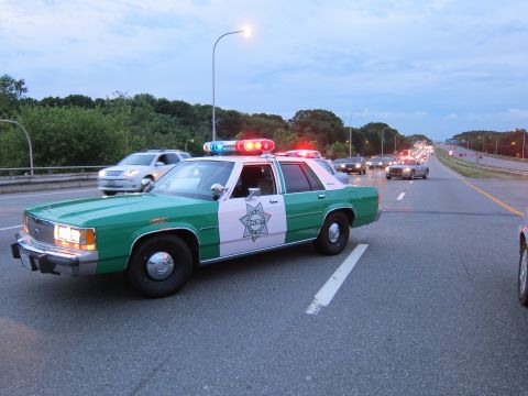 1991 Ford LTD Crown Victoria - green-and-white Ford sheriff sedan runs its light bar on a highway with traffic and a tree line behind it at dusk.