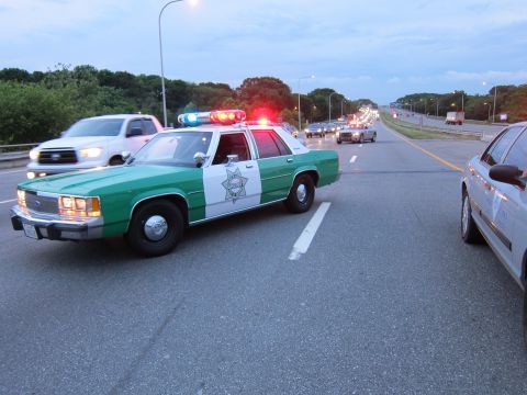 1991 Ford LTD Crown Victoria - green-and-white Ford sheriff sedan runs its emergency lights on a highway as a white pickup passes and traffic backs up in the distance.
