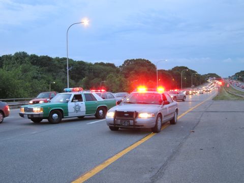 1991 Ford LTD Crown Victoria - green-and-white sheriff sedan and a patrol car with red lights block highway lanes along a yellow centerline as traffic queues behind.
