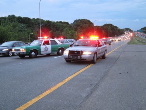 1991 Ford LTD Crown Victoria - green-and-white Ford sheriff sedan and a patrol car run red emergency lights on a divided highway with backed-up traffic at dusk.