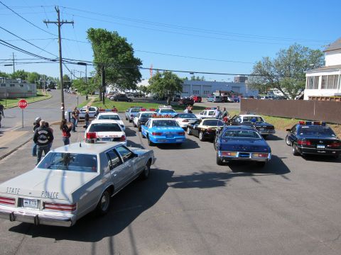 1991 Ford LTD Crown Victoria - parking lot crowded with vintage police cars and motorcycles, led by a silver sedan lettered STATE POLICE near a stop sign.