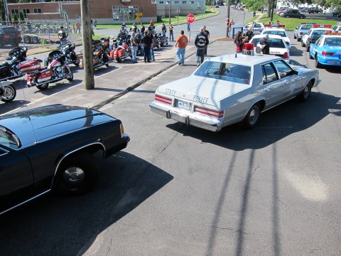 1991 Ford LTD Crown Victoria - silver sedan lettered STATE POLICE drives through a lot lined with parked motorcycles and other vintage police cars while onlookers gather.