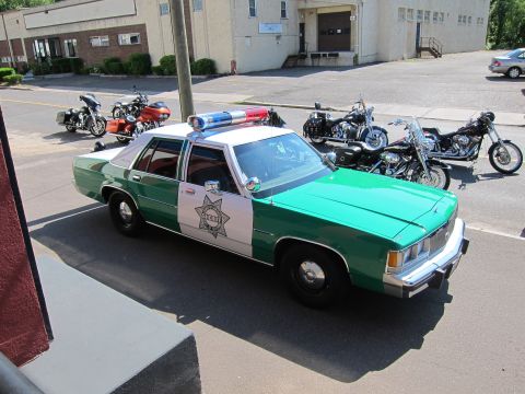 1991 Ford LTD Crown Victoria - green-and-white Ford sheriff sedan with a star emblem on the door sits parked beside a building with several motorcycles lined up behind it.