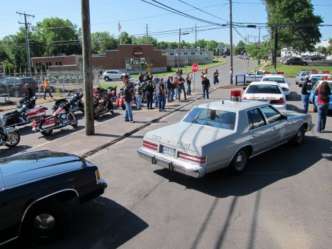 1991 Ford LTD Crown Victoria - silver sedan lettered STATE POLICE sits in a lot among parked motorcycles, a gathered crowd, and other patrol cars near a brick building.