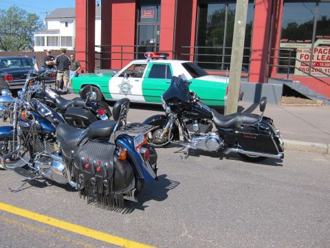 1991 Ford LTD Crown Victoria - Black touring motorcycles are parked along a street with a green-and-white sheriff car and a real-estate sign on a red building behind them.
