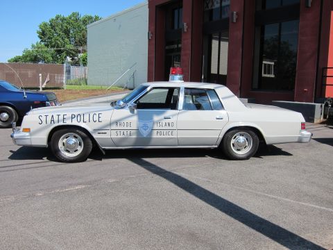 1991 Ford LTD Crown Victoria - white sedan lettered STATE POLICE with a Rhode Island State Police door badge is parked in profile beside a brick building.