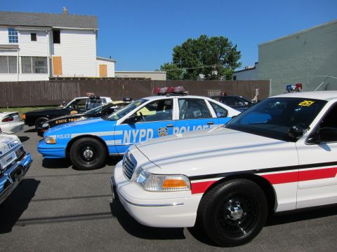1991 Ford LTD Crown Victoria - blue NYPD Ford Crown Victoria, a white sheriff car with a red stripe, and a dark State Trooper car are parked together under a clear sky.