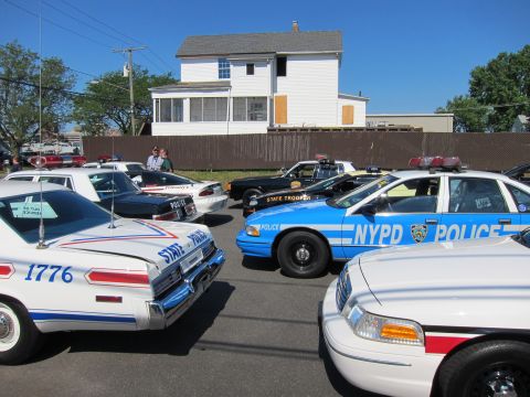 1991 Ford LTD Crown Victoria - cluster of vintage police cars including a blue NYPD Crown Victoria and a white STATE car numbered 1776 is parked before a white house.