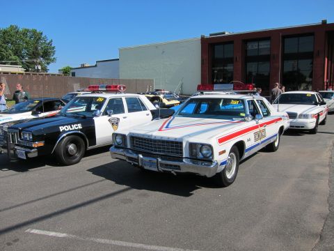 1991 Ford LTD Crown Victoria - white vintage Dodge police car with red and blue stripes parked next to a black 'Police' sedan in front of a brick building.