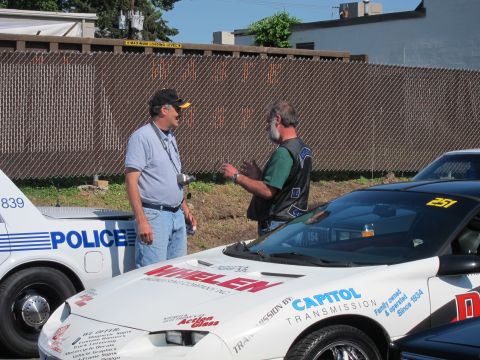 1991 Ford LTD Crown Victoria - Two men converse near a white sports car covered in sponsor decals beside a 'Police' sedan along a chain-link fence.