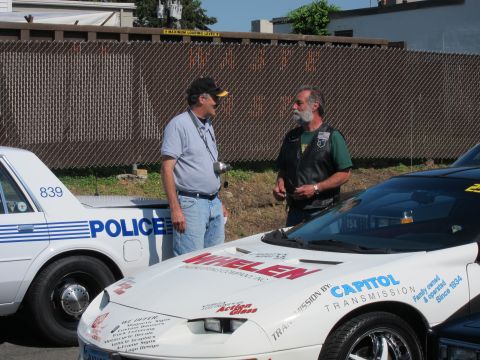 1991 Ford LTD Crown Victoria - Two men stand talking beside a white Pontiac Firebird carrying Whelen and Capitol sponsor decals, next to a 'Police' car.